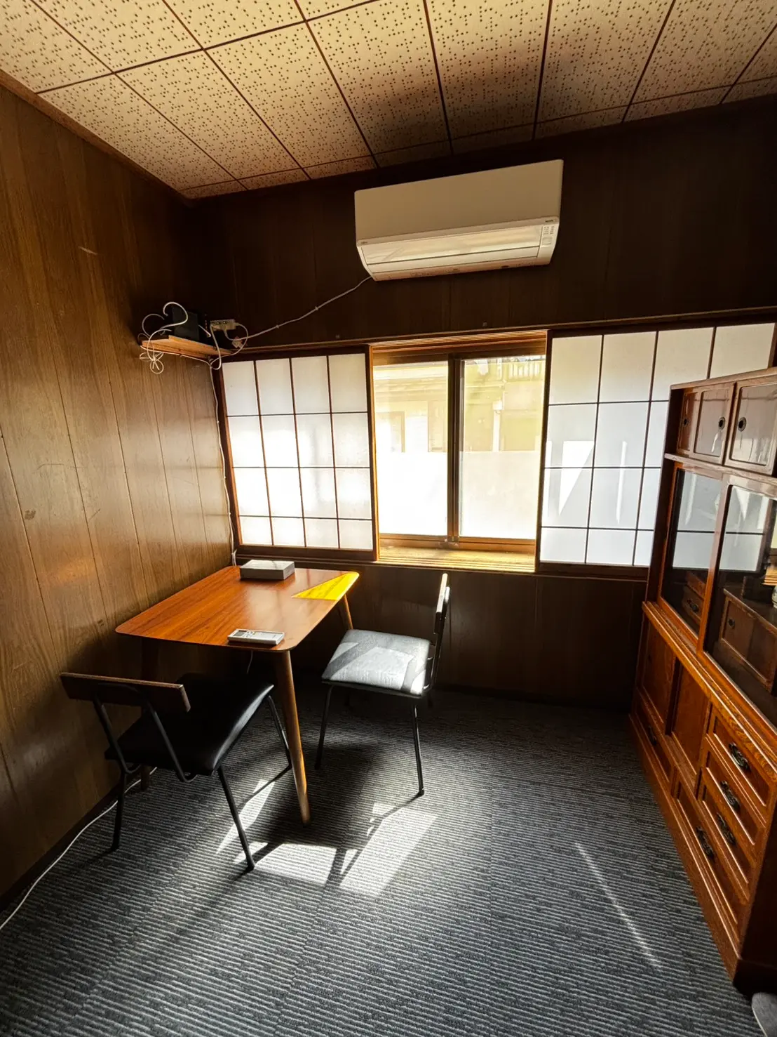 Small study corner with a wooden table, two black chairs, and a window with frosted panels; an air conditioner mounted above the wall.